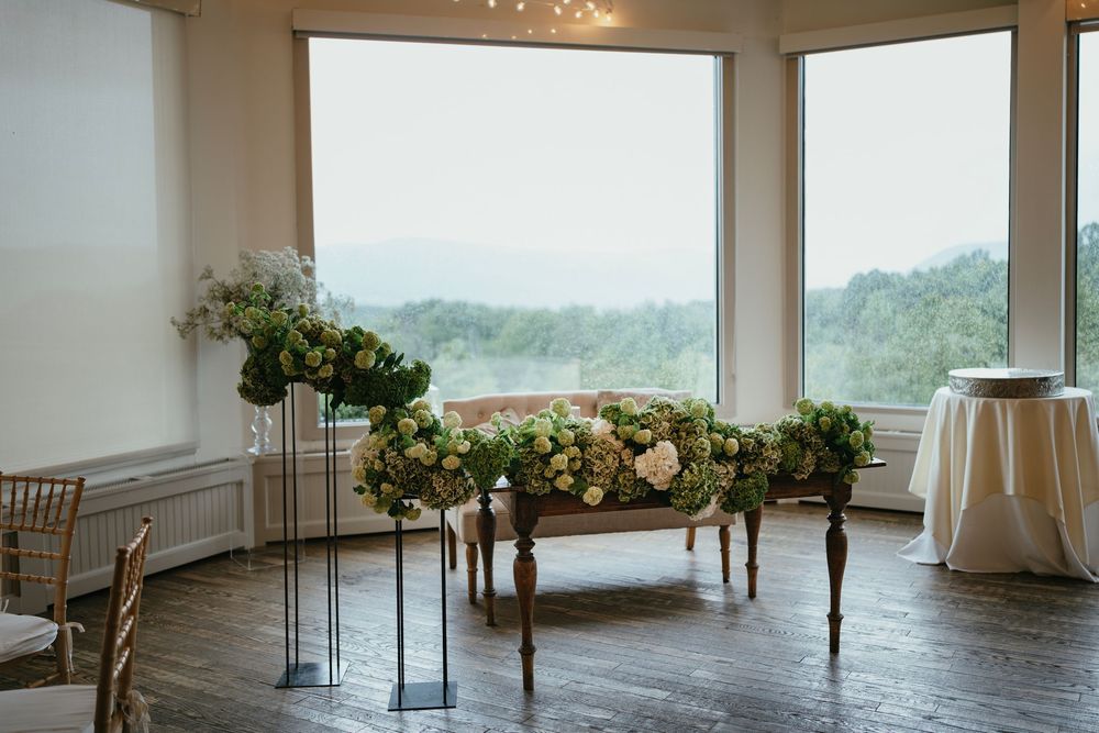 Elegant wedding reception setup with floral arrangements on stands near large windows overlooking a misty landscape.