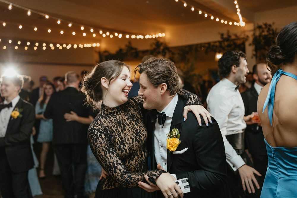 A group of wedding guests dance and celebrate together under warm string lights at an indoor reception venue.