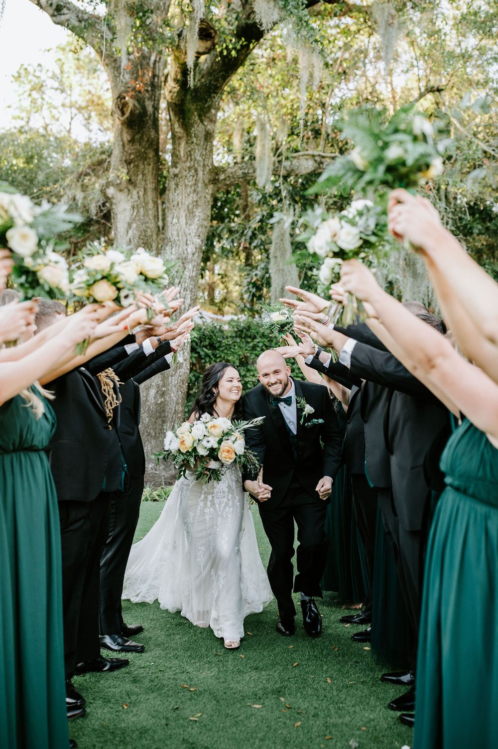 Wedding party stands in a line outdoors with bridesmaids in emerald dresses and groomsmen in black suits.