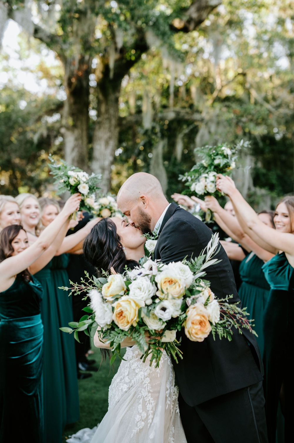 Wedding party in emerald green dresses and black suits pose together outdoors with floral bouquets.
