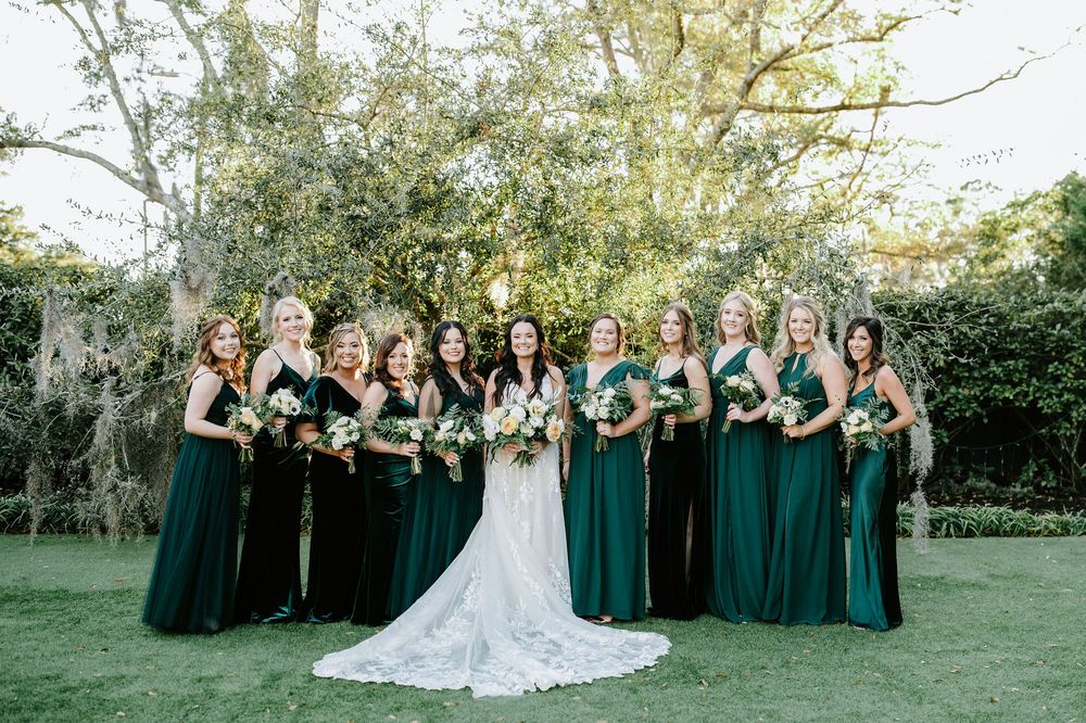 Wedding party in emerald green bridesmaid dresses poses with bride in white lace gown on manicured lawn.