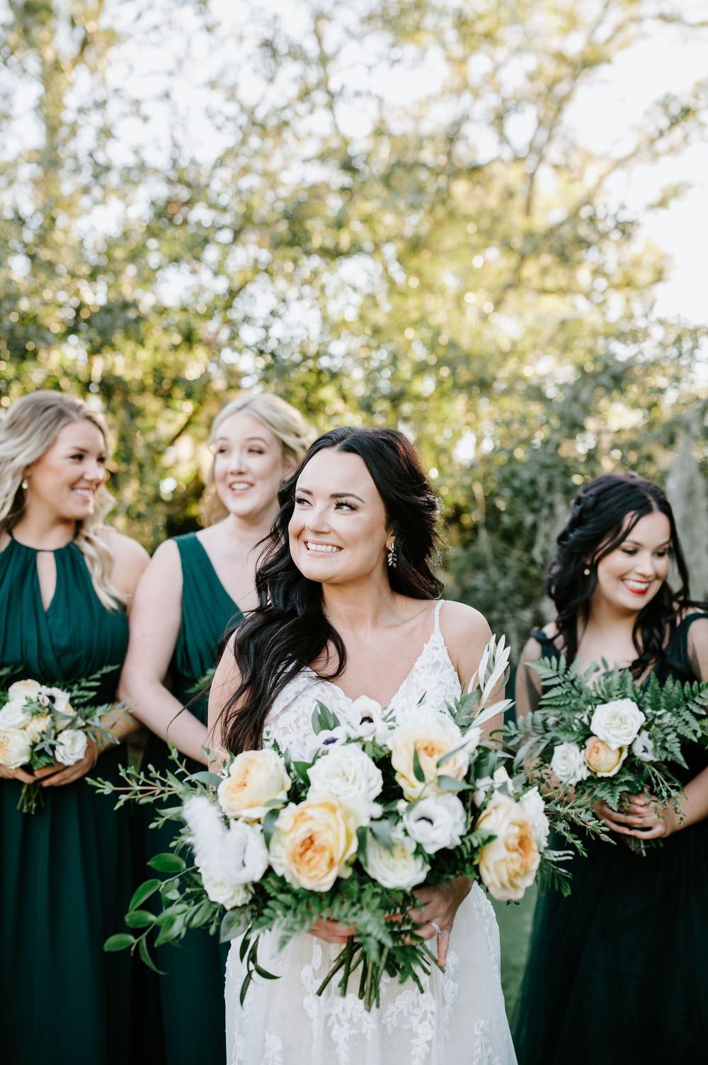 Bridesmaids in emerald green dresses holding yellow and white floral bouquets laugh together in an outdoor setting.