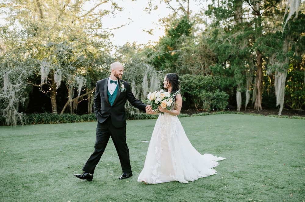 A wedding couple shares a joyful moment twirling and dancing together on a lush green lawn surrounded by trees.