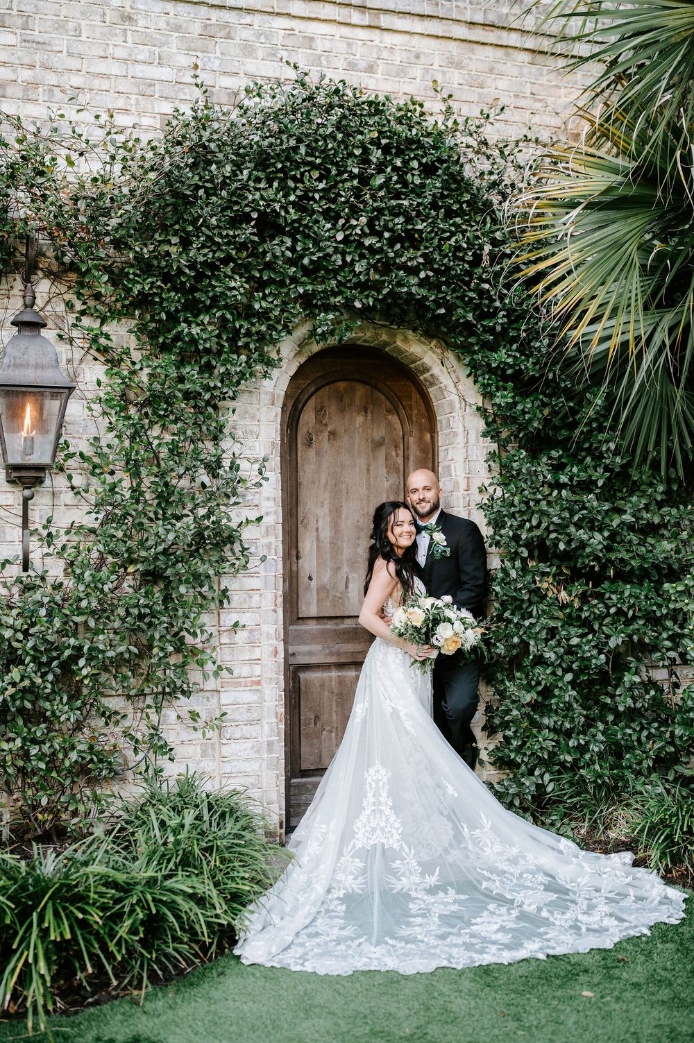 A couple in wedding attire poses against a stone wall with lush green vines and palm fronds at a romantic venue.