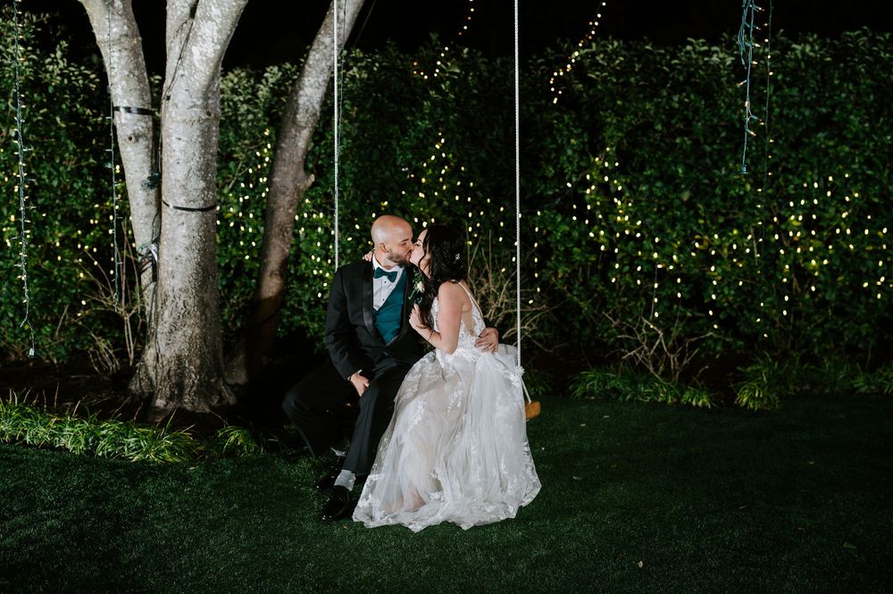 A couple in wedding attire sits together on a forest floor surrounded by illuminated trees and magical string lights.