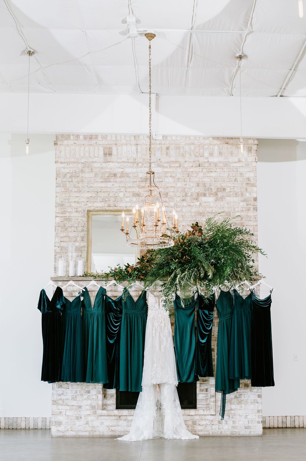 Emerald green bridesmaid dresses and white wedding gown hanging in a row against exposed brick wall with chandelier.