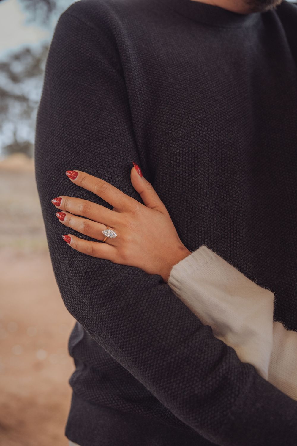 Close-up shots of hands with red nail polish and a sparkling engagement ring against a black sweater.