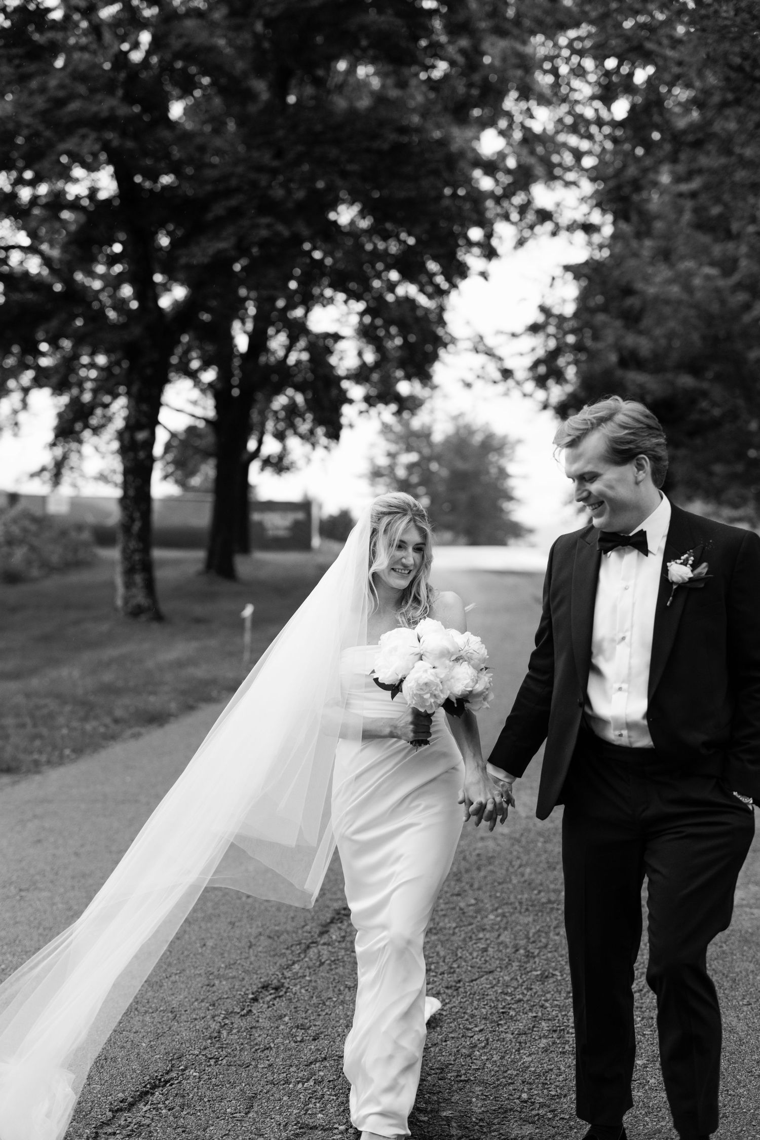 Black and white wedding portrait of couple walking and laughing together in a park setting with trees in background.