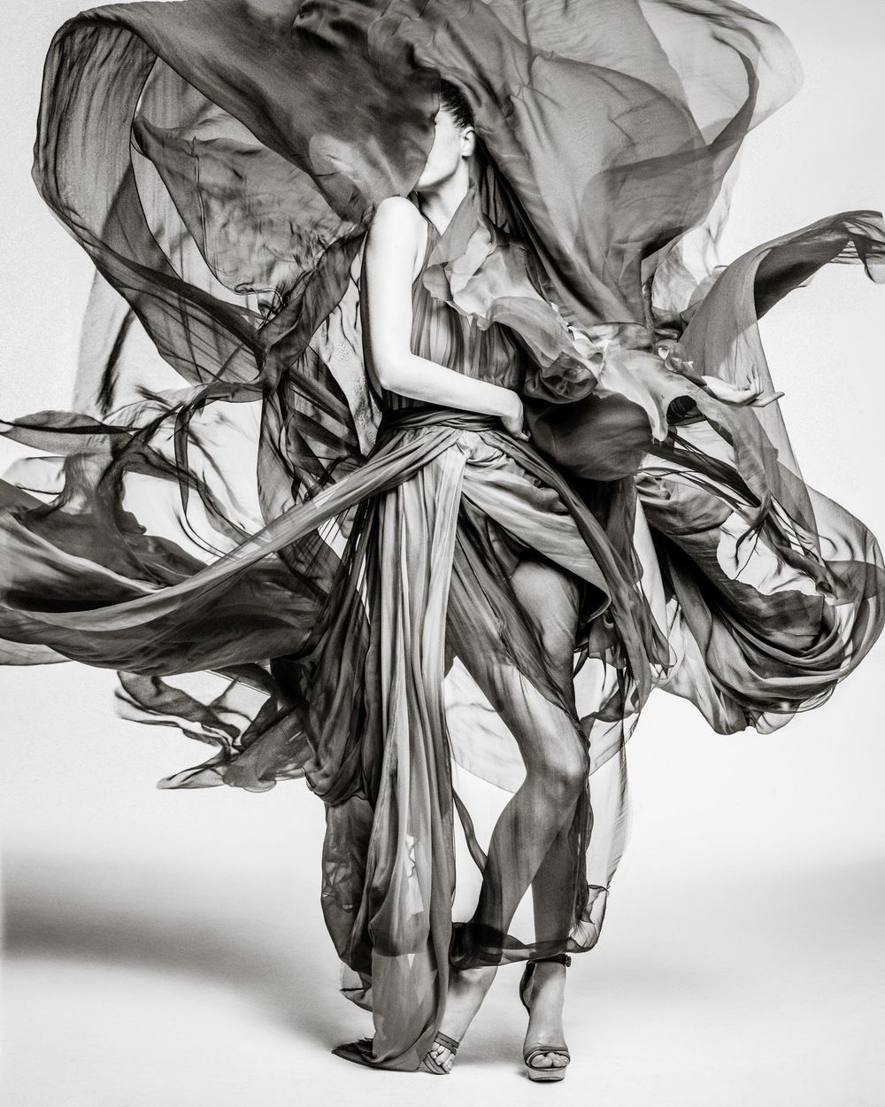 A dramatic black and white fashion photograph showing flowing fabric in motion as a figure poses gracefully.