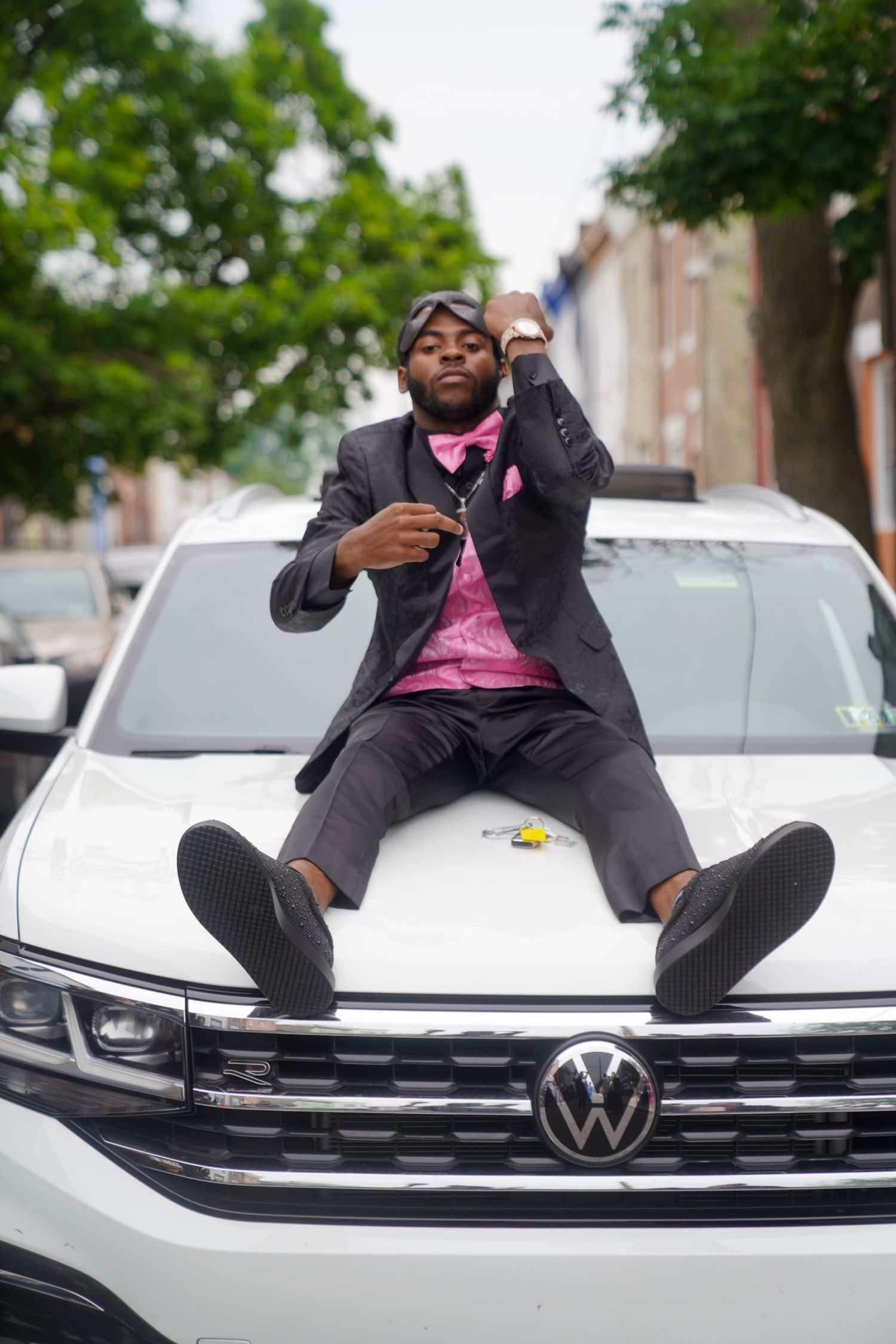 A person in a dark suit with pink accents reclines on the hood of a white Volkswagen car in an urban setting.