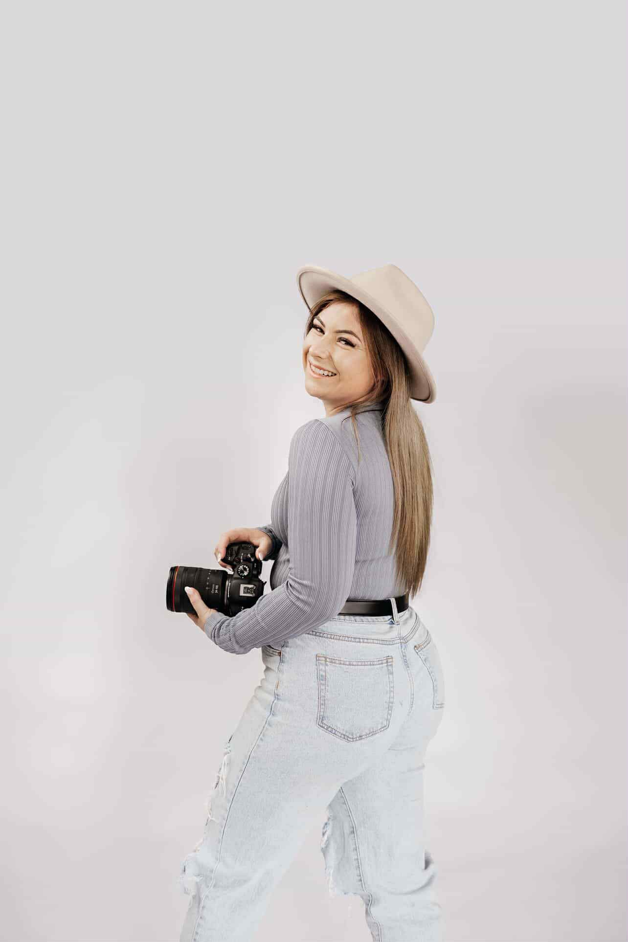 Smiling female photographer in beige hat and jeans holding DSLR camera over shoulder.