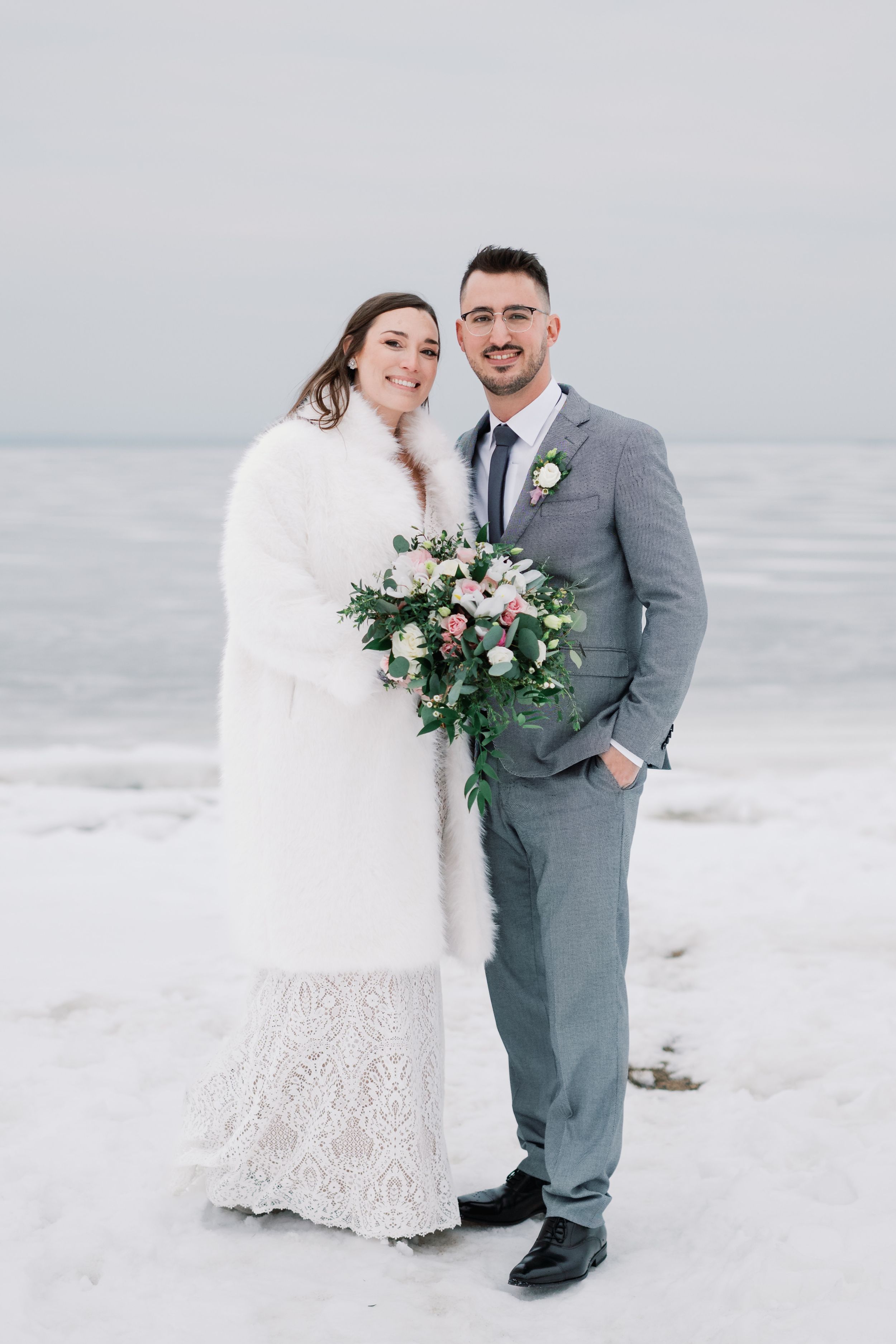 A winter wedding couple poses in the snow, with the bride in a white fur coat holding a pink and green bouquet.