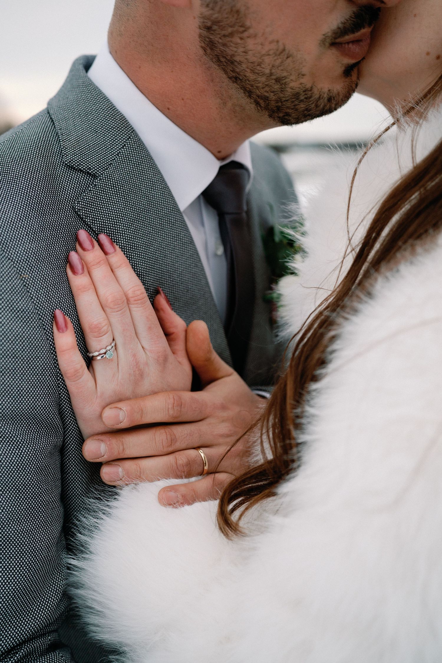 Close-up wedding photo showing hands with rings and a white fur wrap against a gray suit.