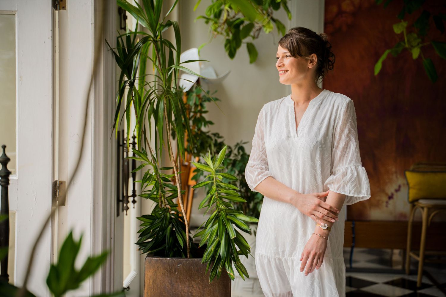 Woman in white dress stands among indoor plants in a bright, sunlit room with modern minimalist decor. Shot by Luminair Studio
