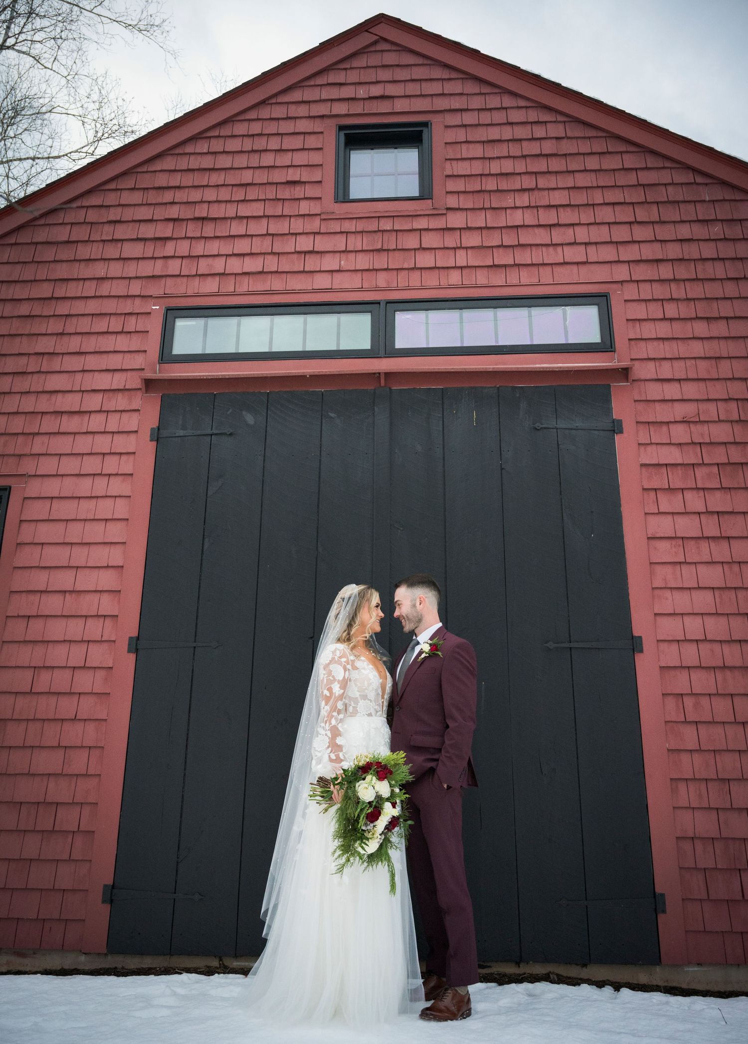 A couple stands in front of a large red barn with black doors on a snowy winter day.