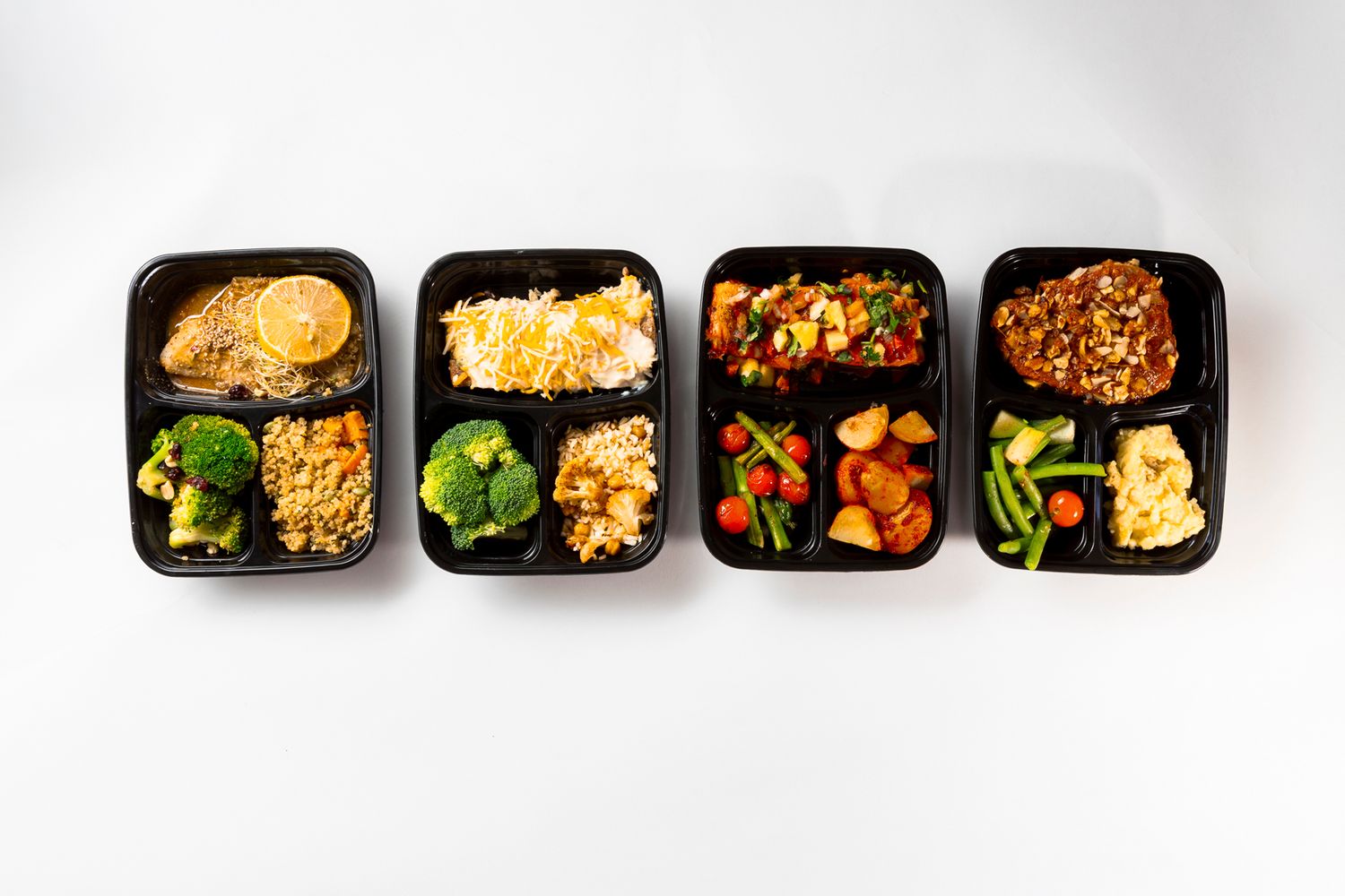 Four black meal prep containers filled with various Asian-inspired dishes including rice, vegetables, and proteins arranged in a row.