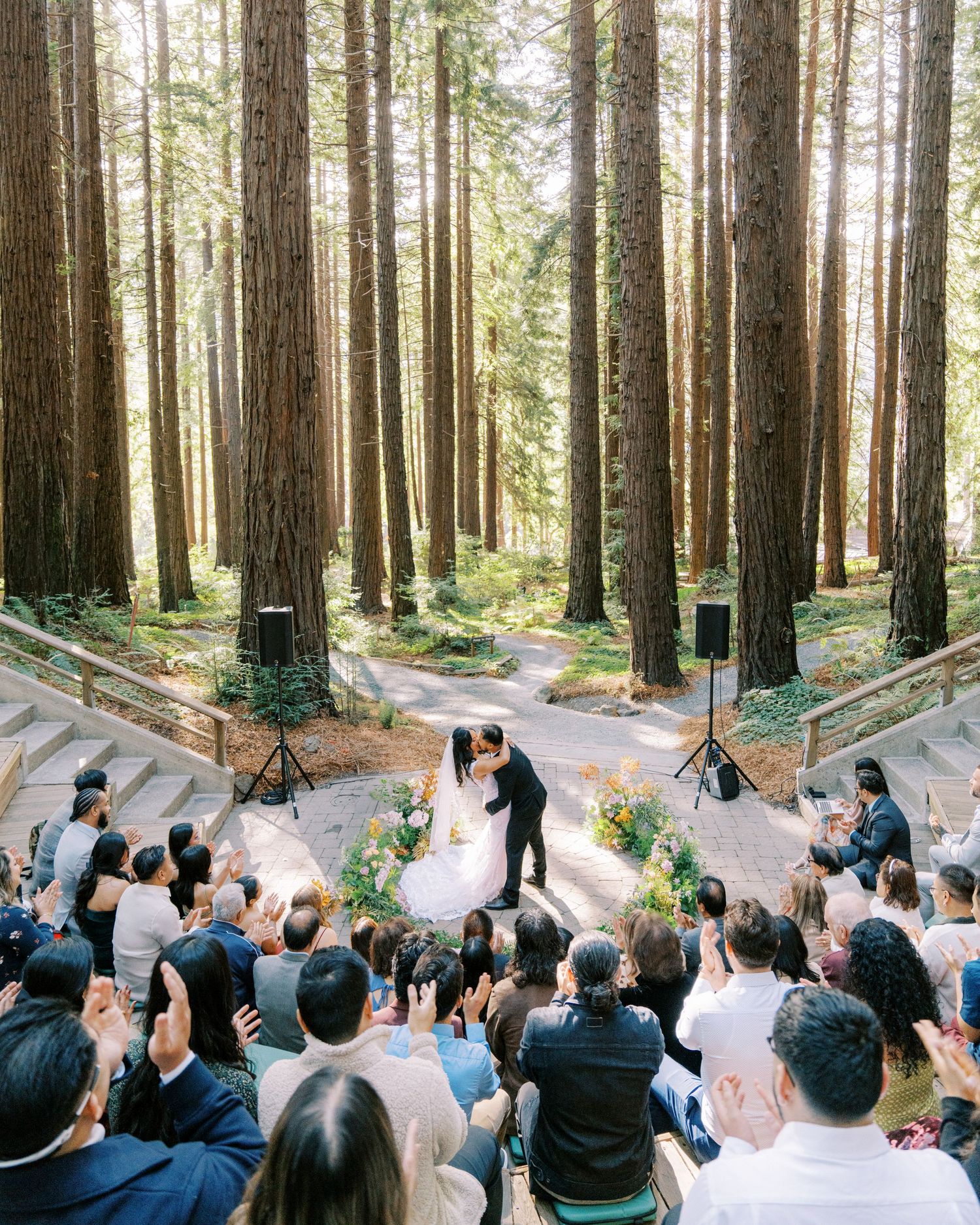 Wedding ceremony in a majestic redwood forest with guests gathered around a floral-lined ceremony space.