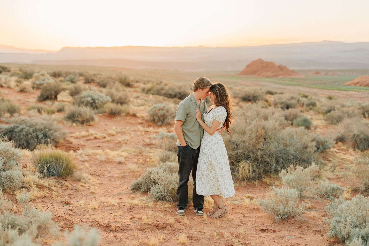 Desert engagement photo session with couple walking among red rock formations and desert brush at sunset.