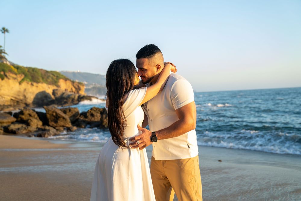 A couple shares a romantic moment on a beach at sunset with rocky cliffs and ocean waves in the background.