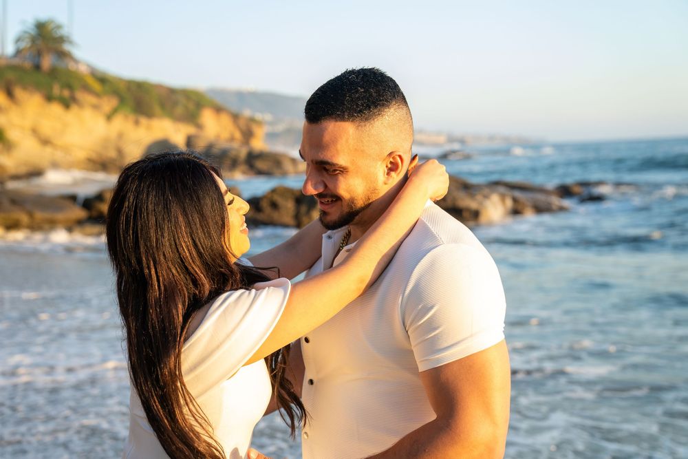 A romantic couple embraces on a rocky beach at sunset with ocean waves and palm trees in the background.