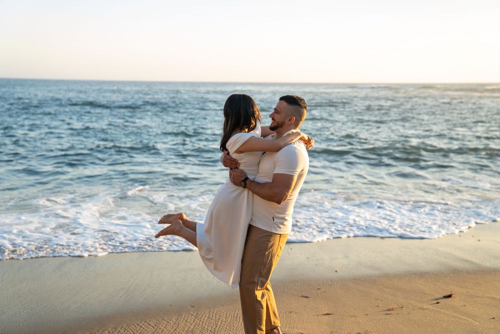 A couple embraces on a sunny beach at sunset with ocean waves rolling in the background.