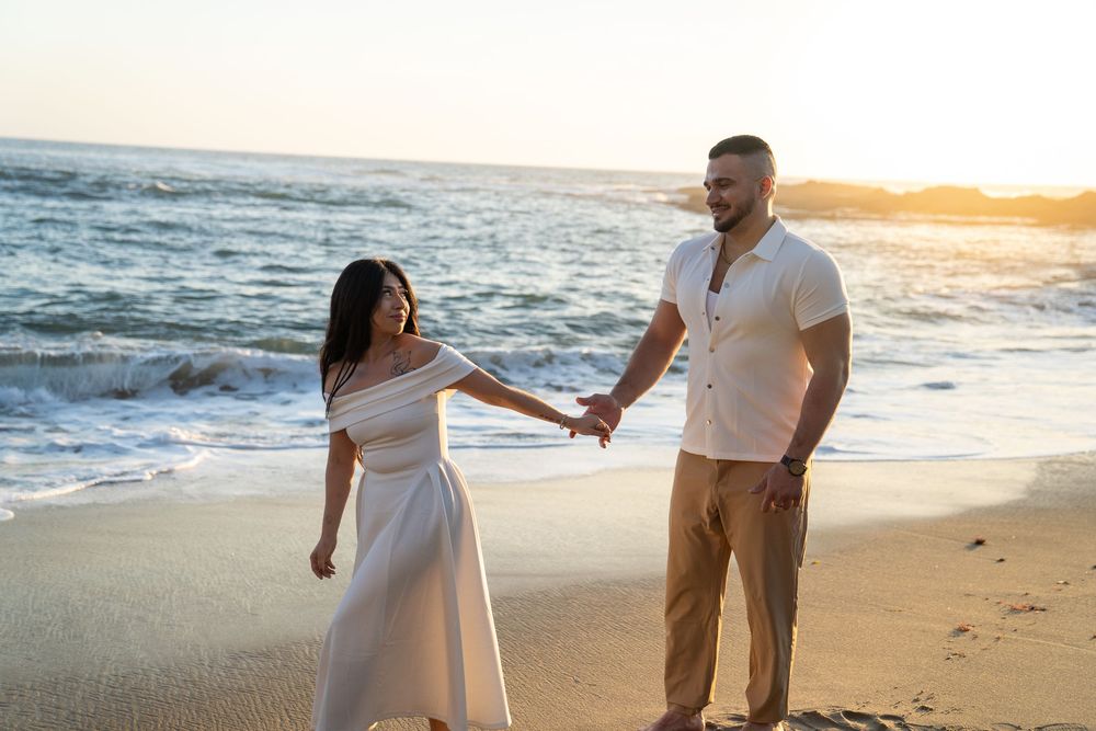 Romantic couple in white attire walking along sandy beach at sunset with ocean waves in background.
