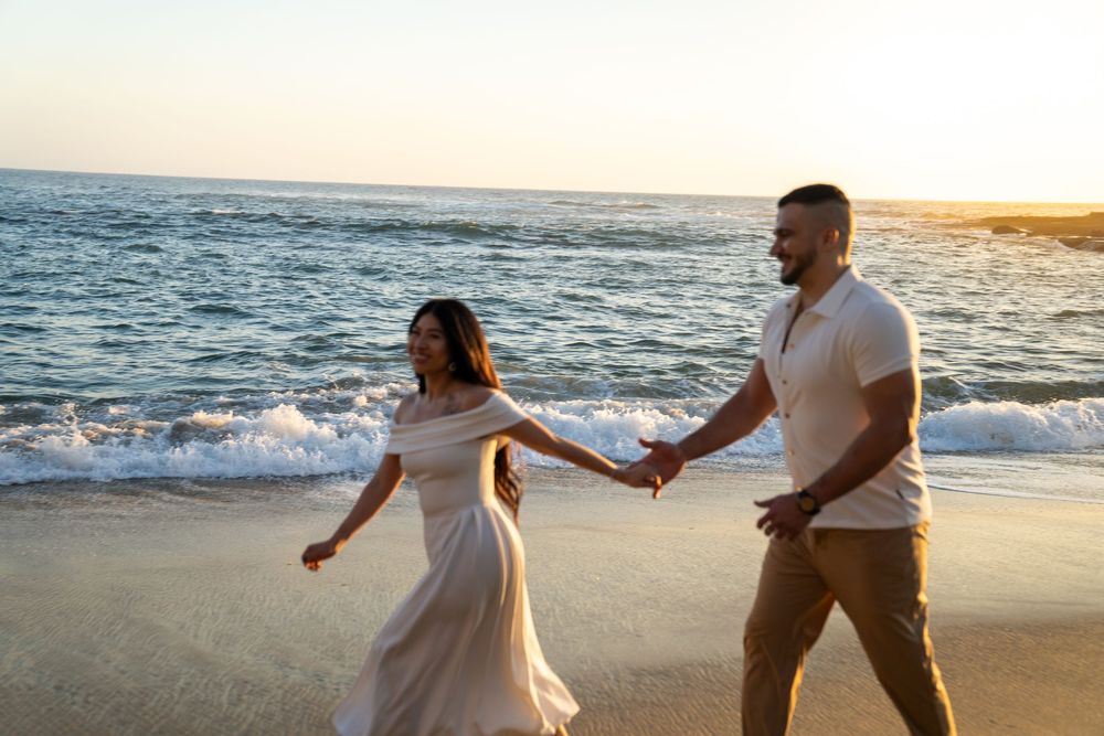 A romantic couple in white outfits holding hands while walking along a sunset beach with crashing waves.
