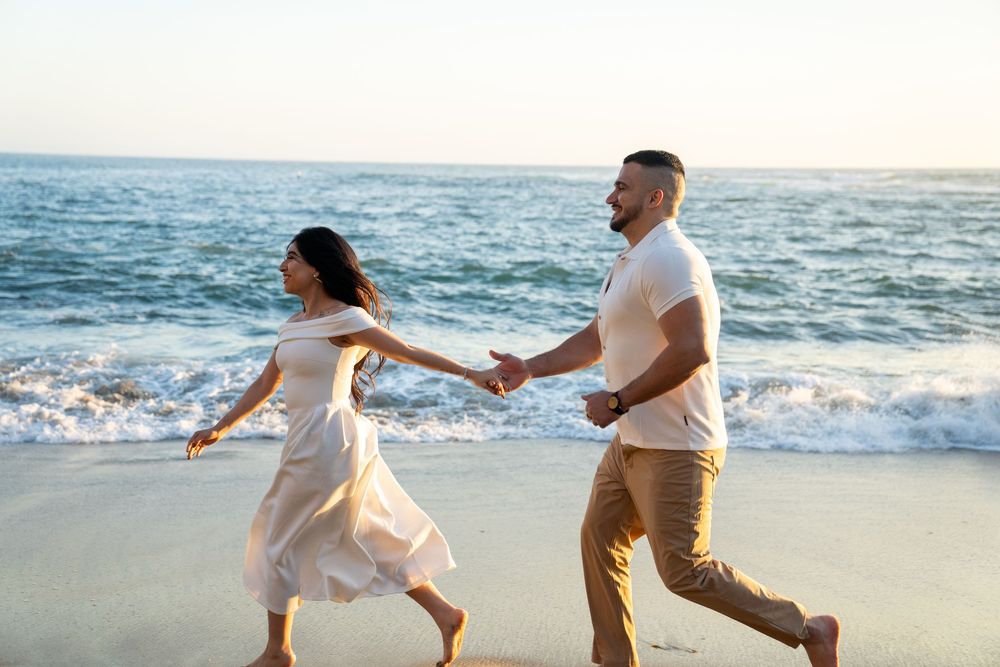 Couple in light colored outfits holding hands and walking along a beach at sunset with waves crashing behind them.