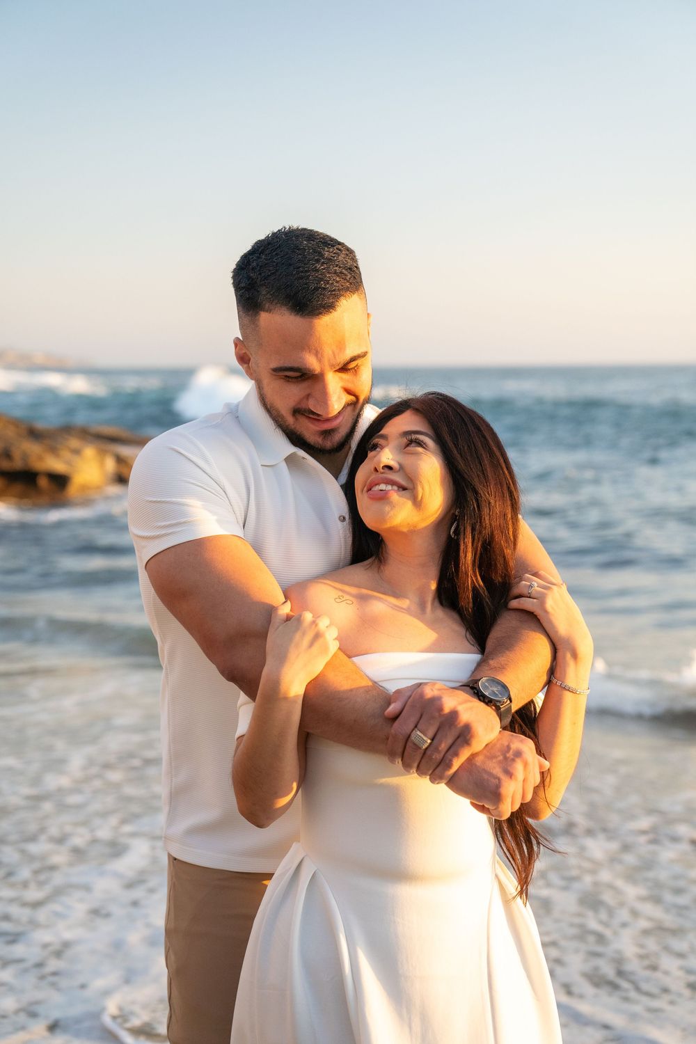 A romantic couple embraces on the beach at sunset with ocean waves and golden light in the background.