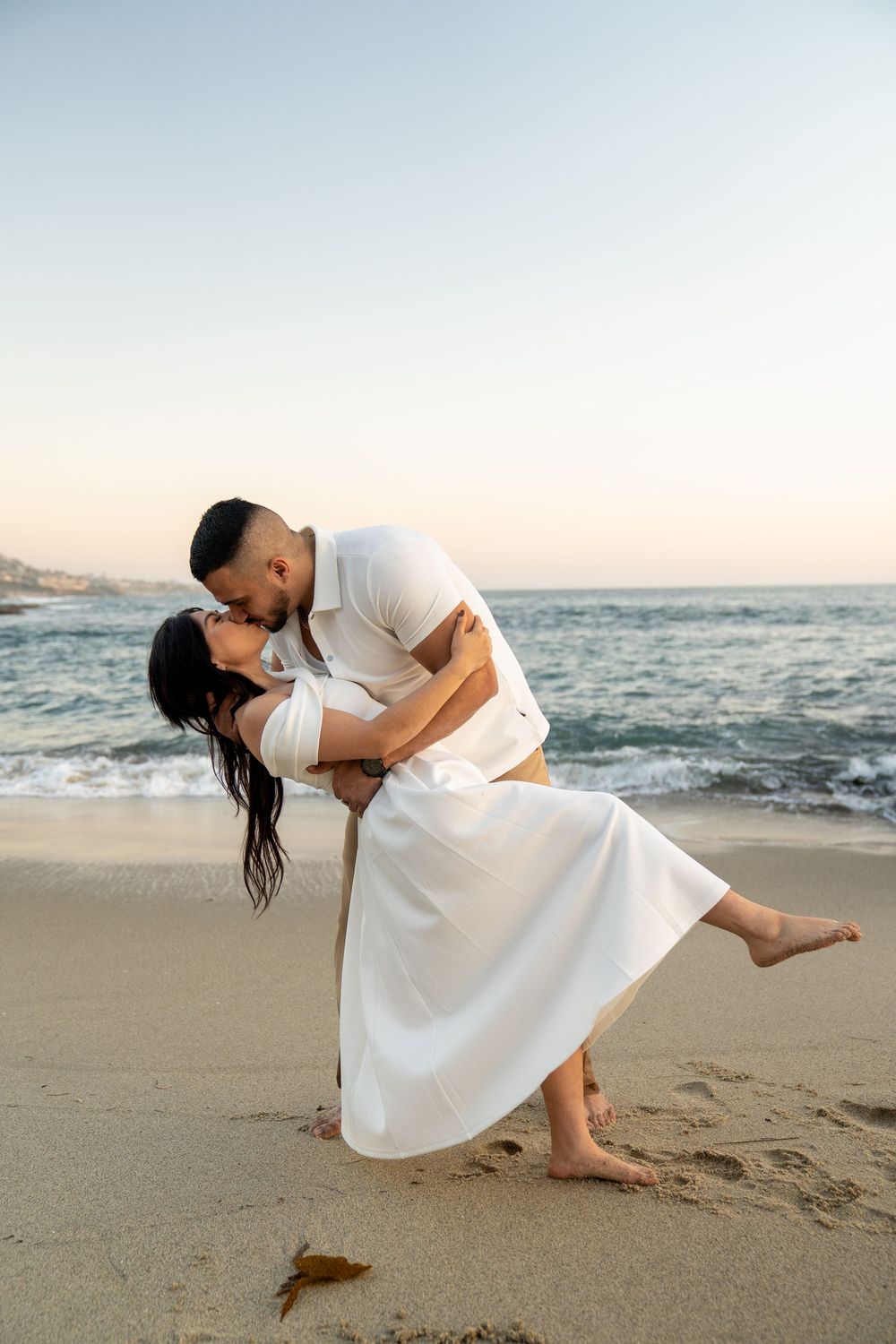 A romantic beach scene with a couple in white embracing in a dramatic dip pose at sunset.