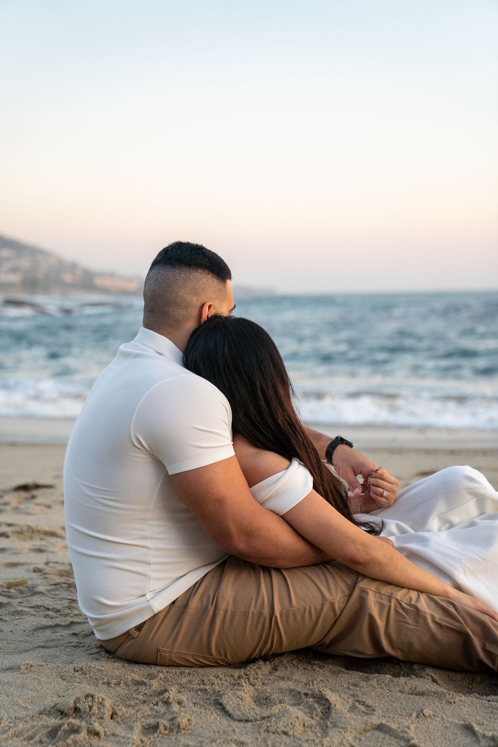 A couple in white clothing sits together on the sand watching the ocean waves at dusk.