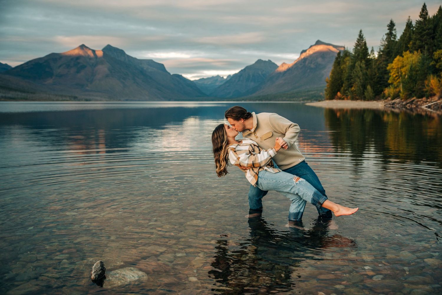 A romantic couple embraces at sunset by a serene mountain lake with evergreen trees and golden light reflecting on the water.