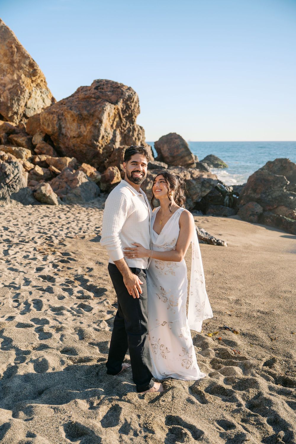 A couple in wedding attire embrace on a scenic beach with rocky cliffs and ocean in the background.