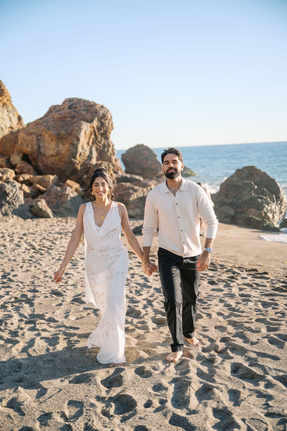 Couple walking and playing together on a sandy beach with large rocks and ocean in the background during sunset.