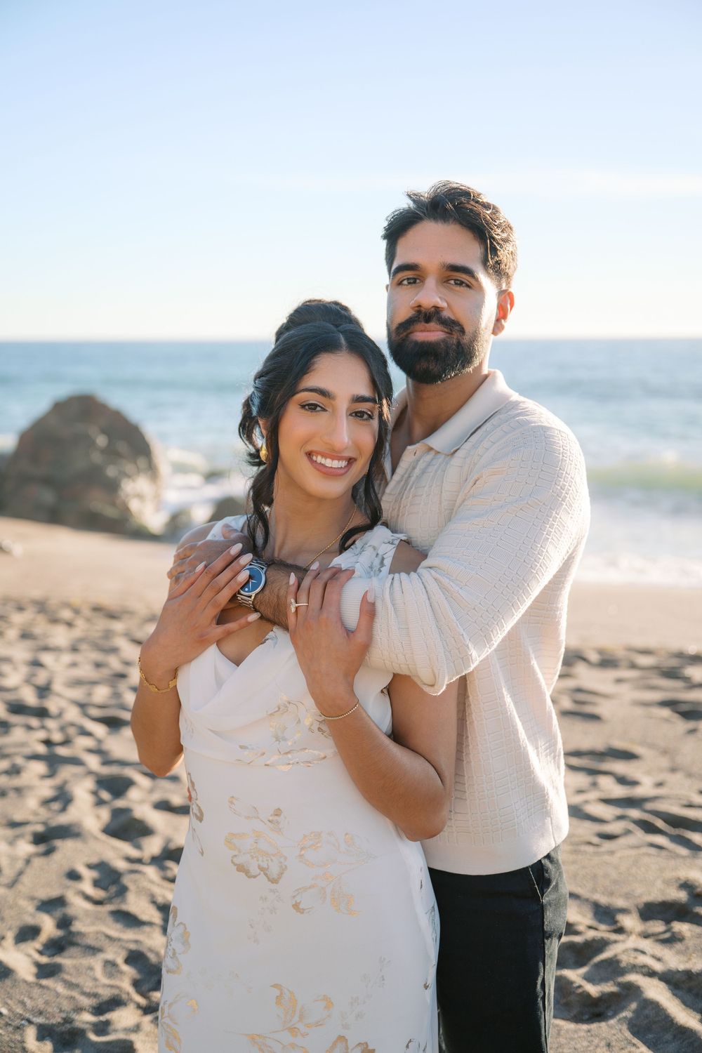 A couple embraces playfully on a sunny beach with waves in the background during a romantic photoshoot.