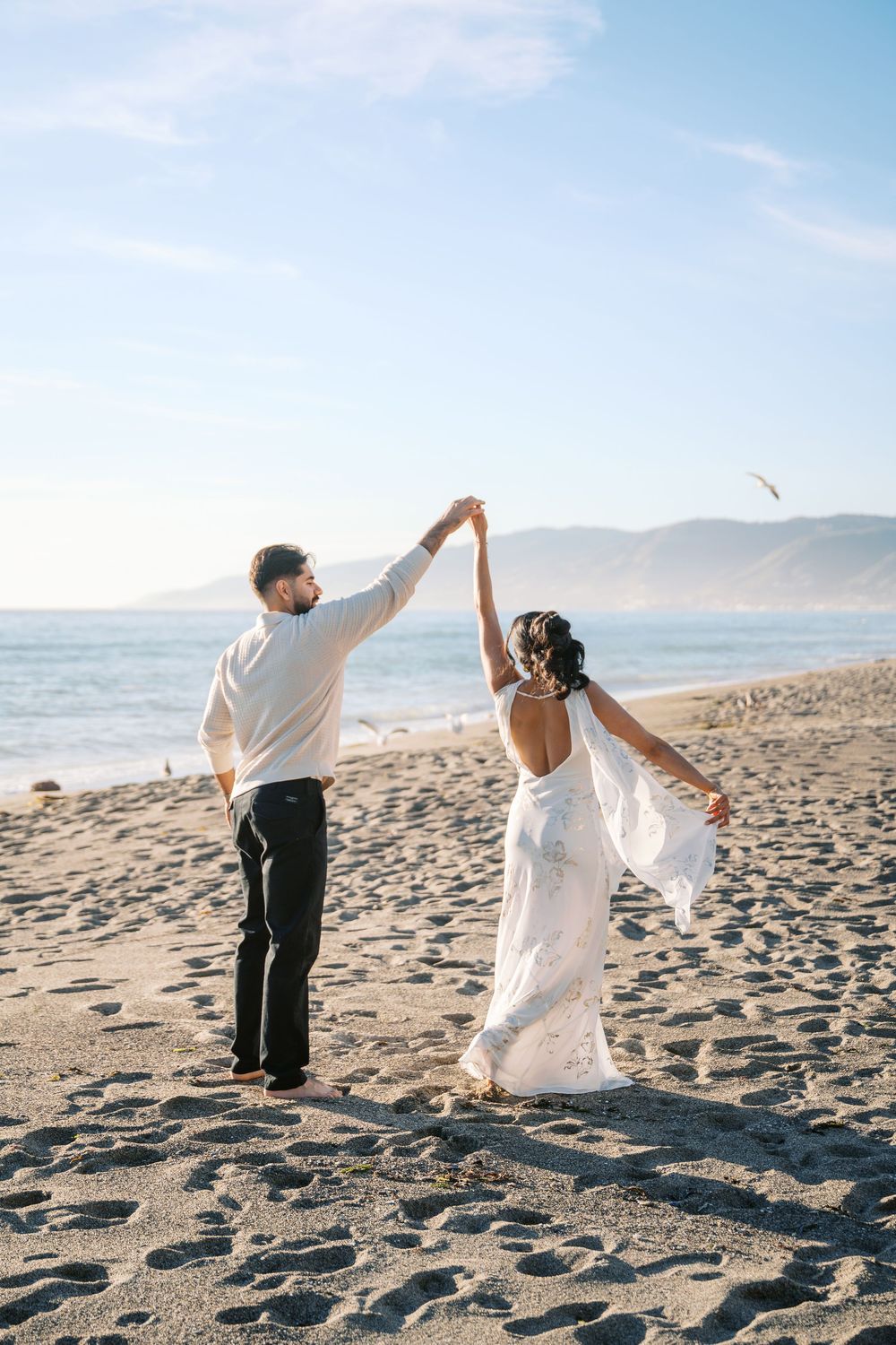 A couple in wedding attire dance together on a sandy beach with mountains and ocean in the background.