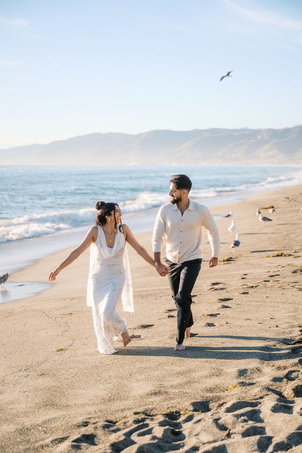 A couple in white clothing runs joyfully along a scenic beach with mountains in the background at sunset.