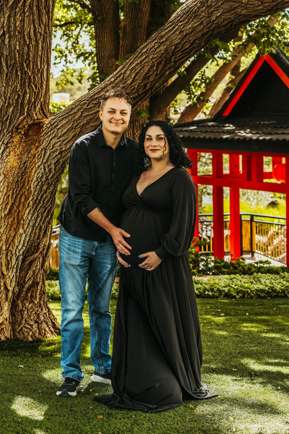 Couple posing for maternity photo shoot in black outfits under large tree with red Japanese garden structure behind them.