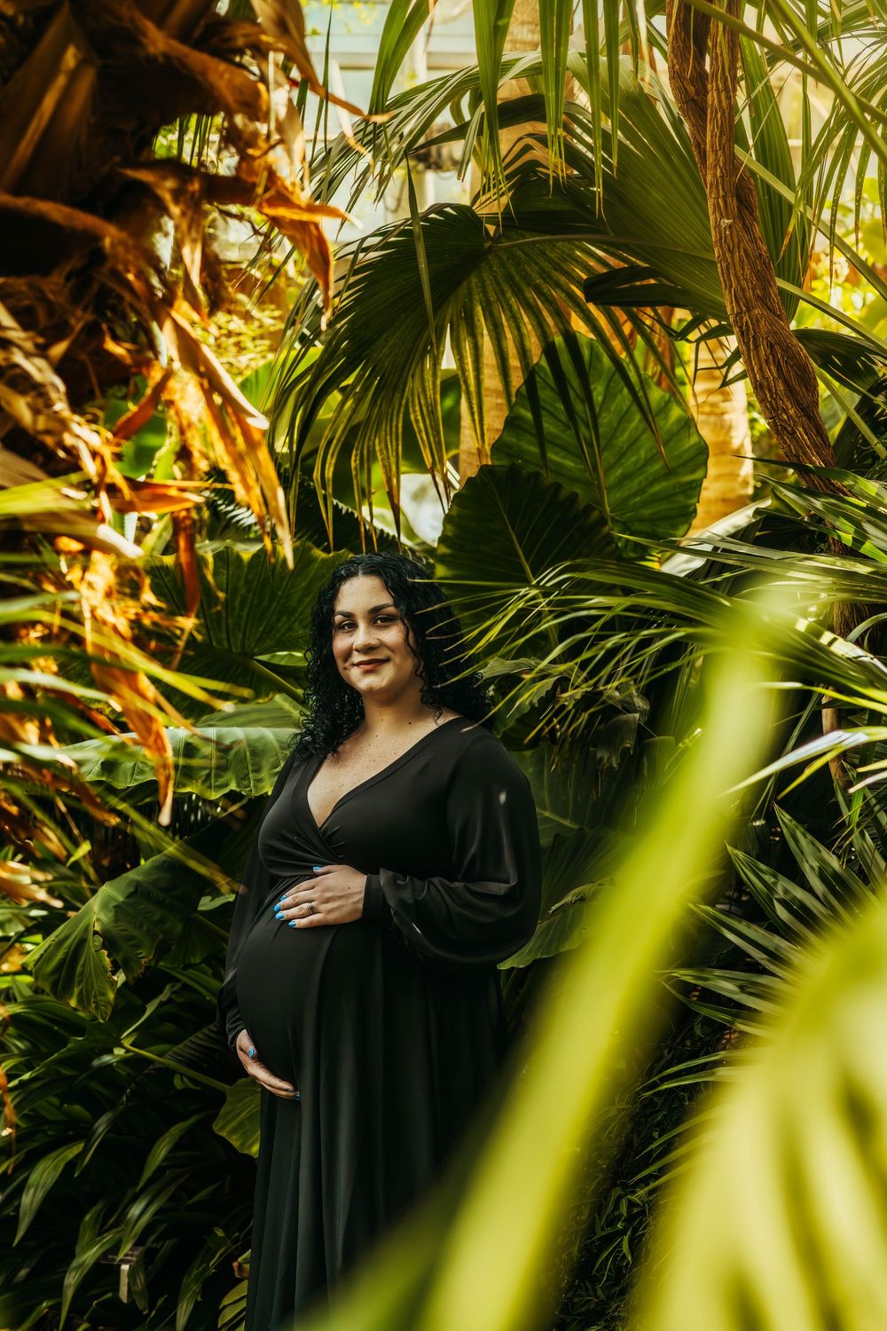 Pregnant woman in black dress posing among lush tropical plants in a greenhouse, cradling her baby bump.