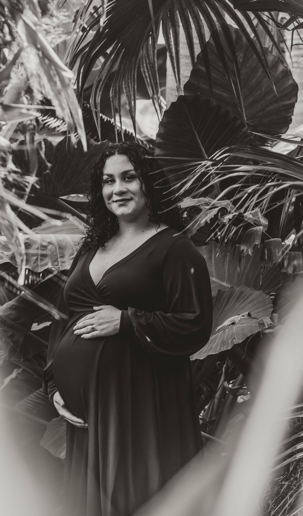 Pregnant woman in black dress cradling baby bump, surrounded by tropical plants in a black and white maternity photo.