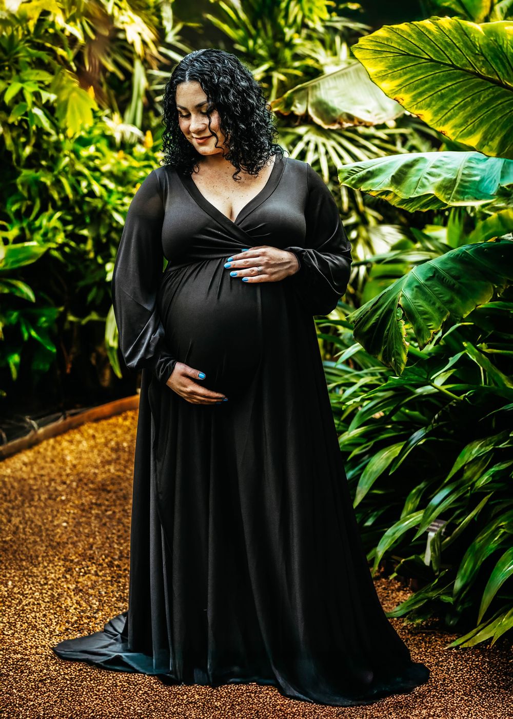 Pregnant woman in elegant black maternity gown poses among lush tropical greenery in a serene outdoor setting.