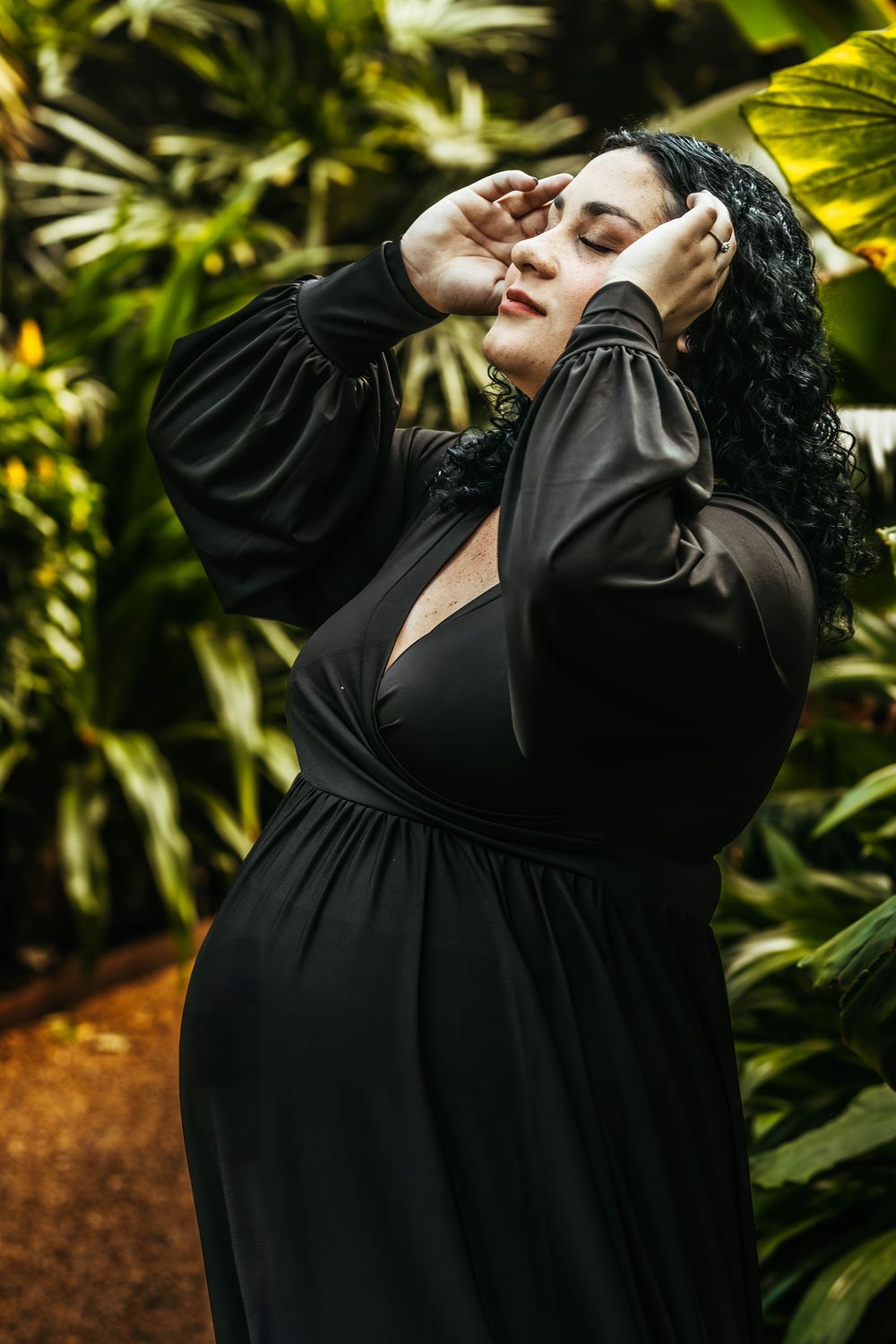 Woman in black wrap dress with curly hair poses gracefully among lush tropical green plants.