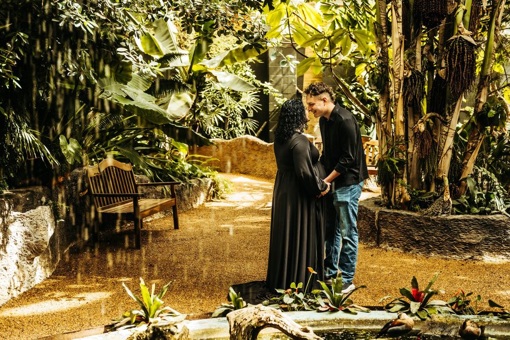Couple sharing a romantic kiss in a lush tropical garden, surrounded by green plants and dappled sunlight.