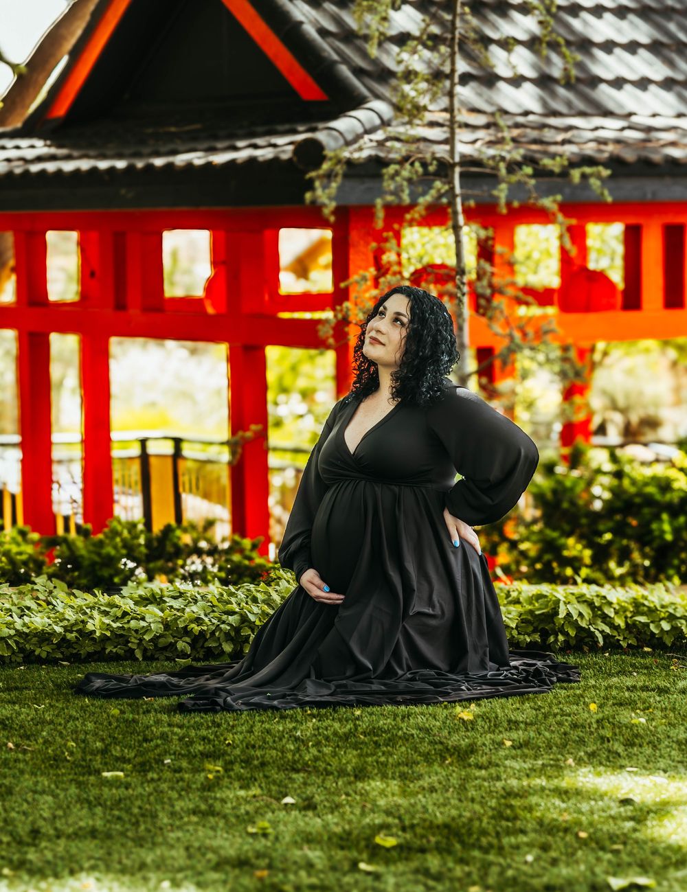 Pregnant woman in black maternity gown kneeling on grass before a red Japanese-style pavilion.