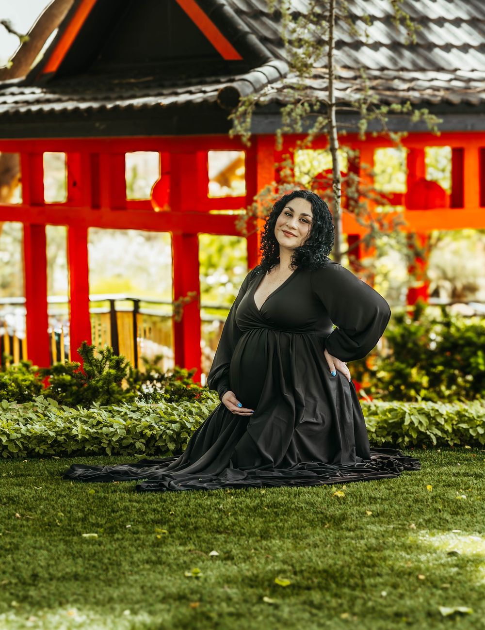 Pregnant woman in black gown poses on grass before red Japanese torii gate in lush garden setting.