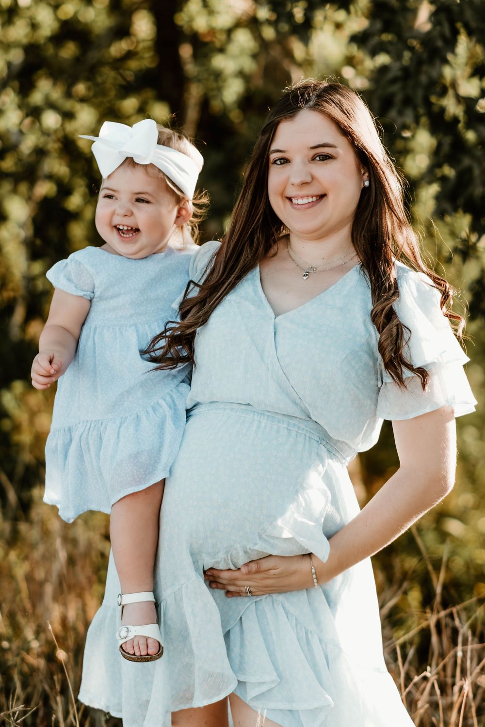 Mother and child in matching light blue dresses pose together outdoors in sunlit garden setting.