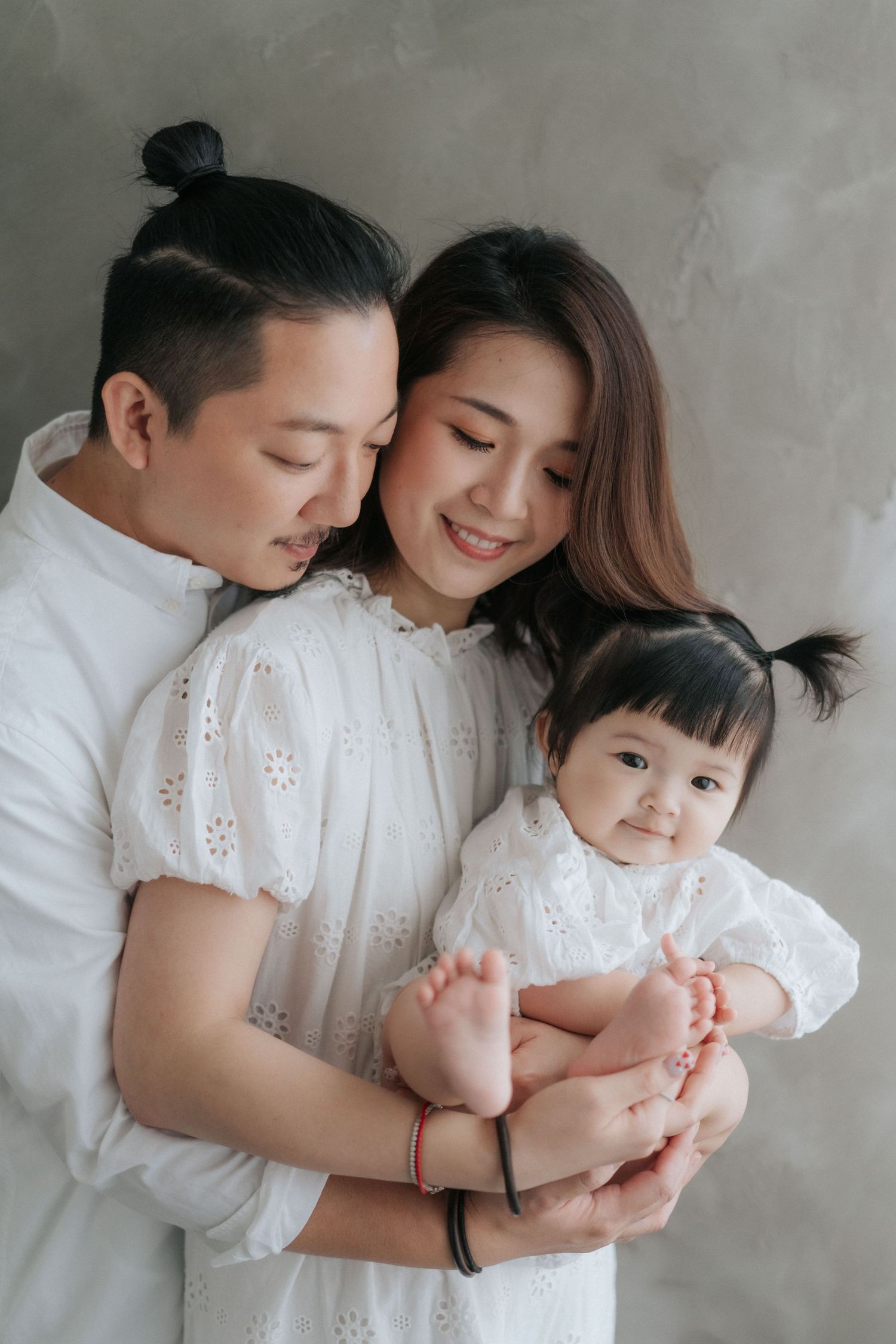 Asian family portrait with parents in white holding their baby girl, smiling warmly against a gray background.亞洲家庭合影，父母穿著白色衣服，抱著他們的女嬰，在灰色背景下
