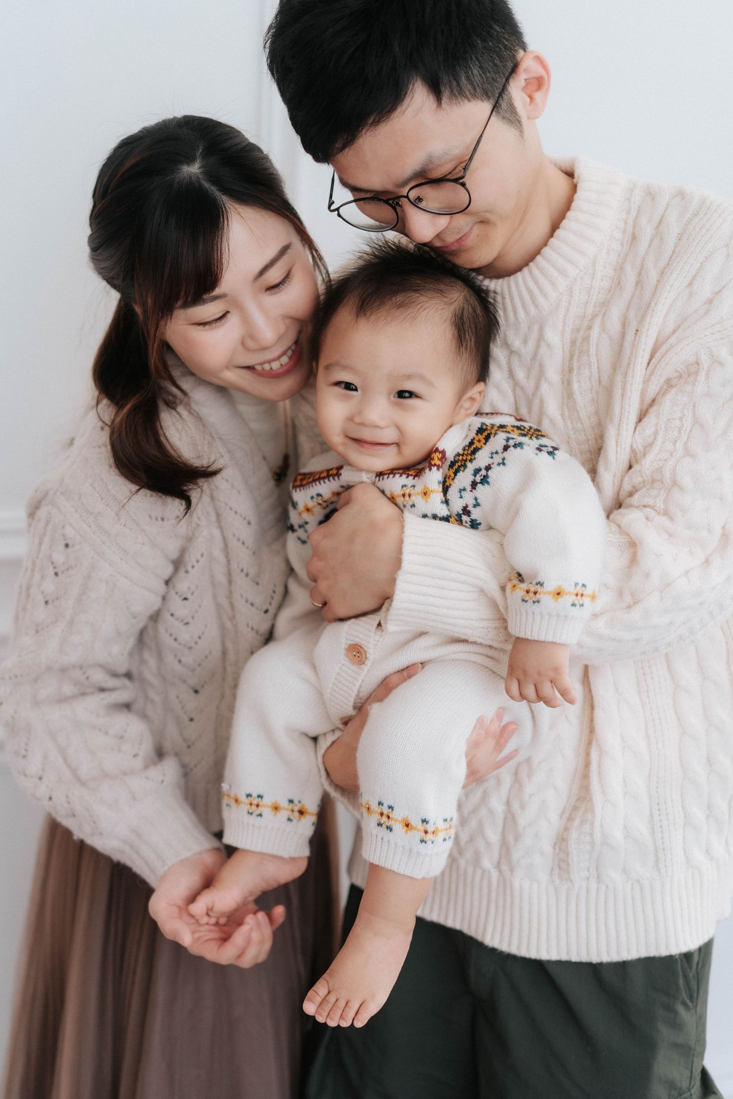 A family in white and gray casual wear lies together on a light background showing tender moments with their baby.一家人穿著白色和灰色休閒裝，躺在淺色背景上