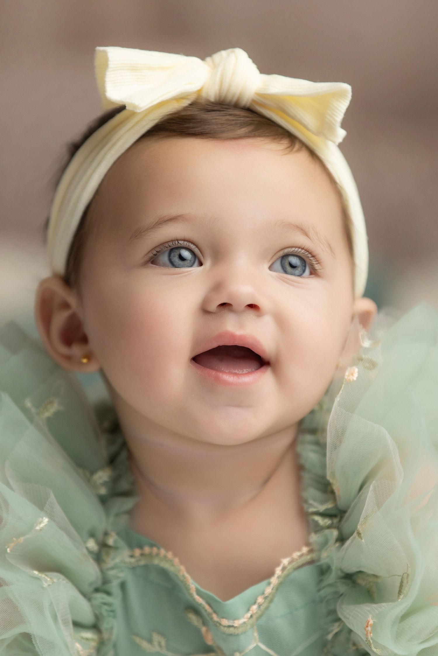 Baby wearing a cream bow headband and mint green tulle dress smiles while looking up.