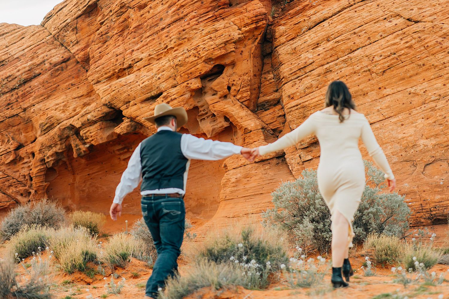 Couple in casual attire poses together along dramatic red sandstone cliffs in the desert.